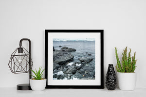 Rocky Antarctic shoreline with restrained grey-blue colour and a broad glacier on the horizon.