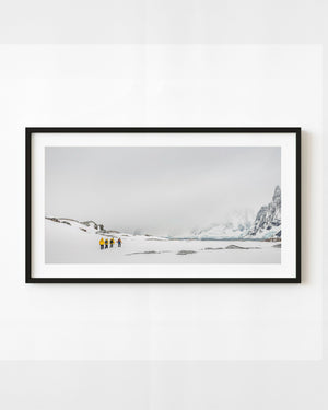 Hikers in yellow jackets cross an Antarctic snowfield beneath mountain cliffs fading into mist.