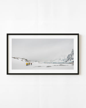 Hikers in yellow jackets cross an Antarctic snowfield beneath mountain cliffs fading into mist.