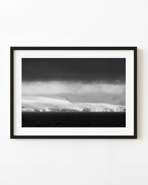 Bright Antarctic ice cliffs beneath dark storm clouds and blowing snow above the ocean.