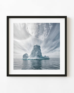 A pale blue Antarctic iceberg rises from grey-blue water beneath sweeping bands of cloud.