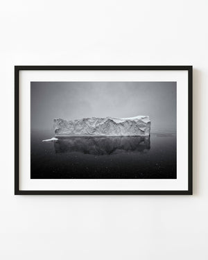 A broad Antarctic iceberg reflects in dark calm water during snowfall in black and white.