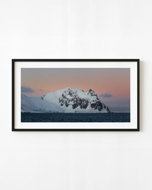 A snow-covered Antarctic mountain rises above dark blue sea beneath a pink dusk sky.