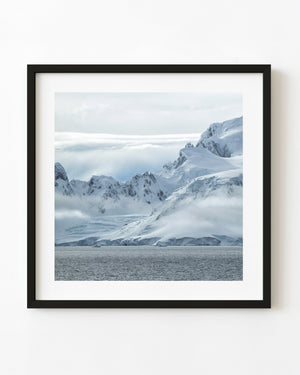 Snow-covered Antarctic mountain ridges beneath low clouds above grey water.