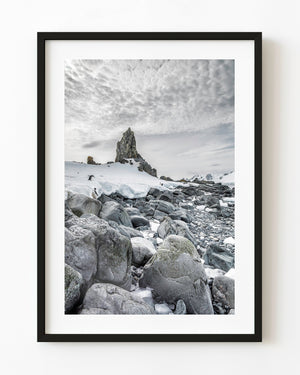 Antarctic shoreline boulders beneath a snowy pinnacle and cloudy sky.
