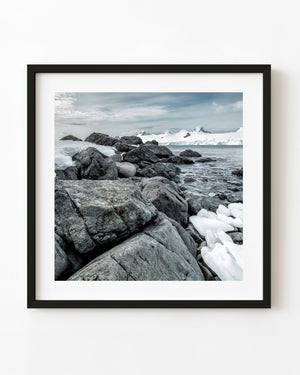 Dark Antarctic shoreline boulders beneath distant snowy peaks.