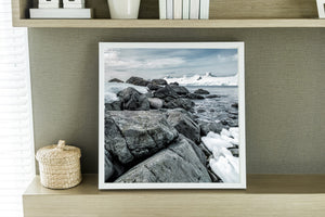 Square Antarctic coastal photograph with stones, sea and distant peaks.