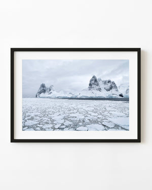 Broken Antarctic sea ice beneath jagged mountain peaks and grey clouds.