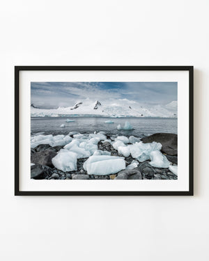 White Antarctic shore ice on a dark pebble beach beneath distant mountains.