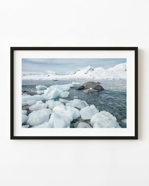 Pale Antarctic shoreline ice beside shallow water and snowy mountains.