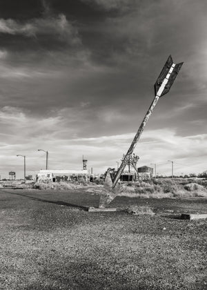 Leaning arrow sculpture beside low roadside buildings beneath a cloudy Arizona sky.