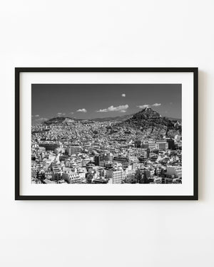 Dense Athens rooftops spread beneath rocky hills and scattered clouds in black and white.