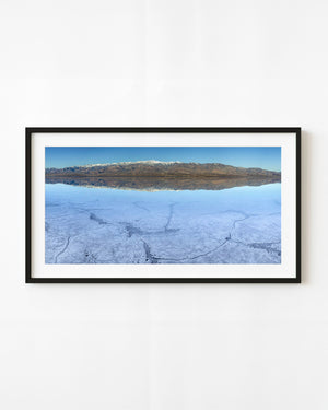 Mountains and blue sky reflect in still Badwater Basin water above patterned salt in Death Valley.