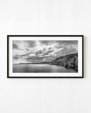Dramatic clouds gather above Ballintoy coastal cliffs and calm sea in black and white.