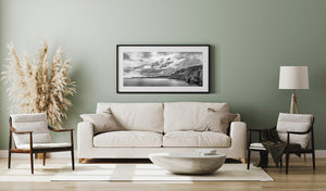 Panoramic monochrome coastal photograph with rugged cliffs, calm sea and textured storm clouds.