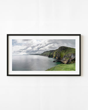 Green-topped Ballintoy cliffs recede beside a quiet silver sea beneath layered clouds in Northern Ireland.