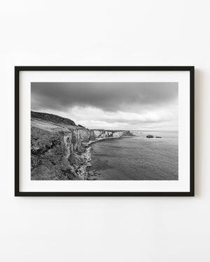 Pale Ballintoy cliffs curve beside open water beneath dark clouds in black and white.
