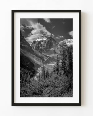 A snowcapped Banff peak rises above a rocky valley and dark conifers in black and white.