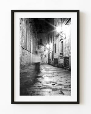 A blurred figure passes through a Barcelona night laneway beneath starburst lamps in black and white.