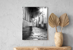 Black and white Barcelona street photograph with a passing figure and wet cobblestone reflections.