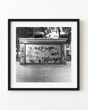 A closed Barcelona newsstand has a graffiti-covered shutter beneath trees in black and white.