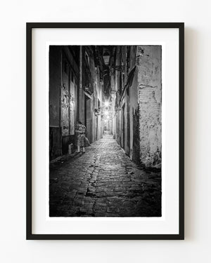 A woman stands beside a doorway in a narrow Barcelona night lane lit by starburst streetlights.