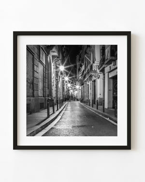 Starburst lamps illuminate a narrow Barcelona street between shuttered shops and balconies in black and white.