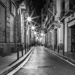 A reflective roadway recedes between close Barcelona facades beneath a sequence of bright night lamps.