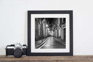 Square monochrome Barcelona street view with closed shutters, reflective paving and dark upper facades.