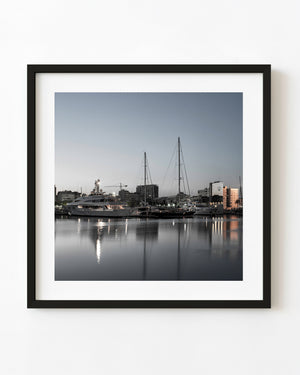 Yachts and tall masts reflect in calm Port Vell marina water beneath a pale Barcelona dawn sky.
