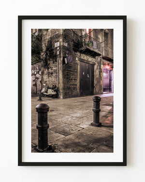 Warm light reveals a weathered Barcelona stone corner with a parked scooter and foreground bollards at night.