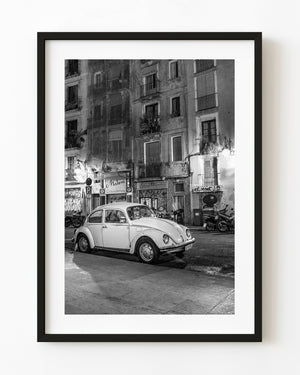 A white Volkswagen Beetle is parked beneath Barcelona balconies and dark windows at night in black and white.