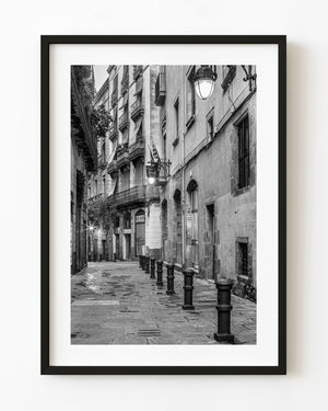 Bollards line a wet Barcelona stone street beneath balconies and glowing lamps in black and white.