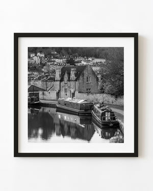 Moored narrowboats reflect in still Bath canal water beneath stone houses and hillside buildings in black and white.