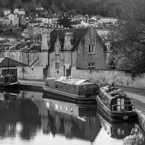Two narrowboats sit beside a stone towpath with calm reflections below Bath’s waterside houses.