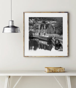 Stone houses and a retaining wall rise beyond reflected boat hulls beneath a wooded hillside.