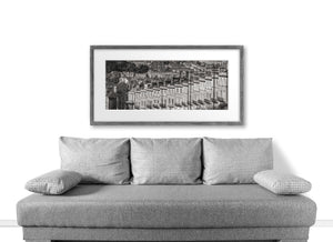 Sofá gris con cojines a cuadros frente a un paisaje urbano en blanco y negro enmarcado sobre una pared blanca.