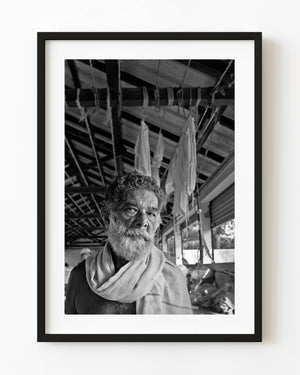 Black and white portrait of a bearded Indian man beneath workshop ropes and roof beams.