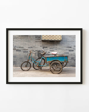 A turquoise cargo tricycle and bicycle stand against a grey brick wall in Beijing.