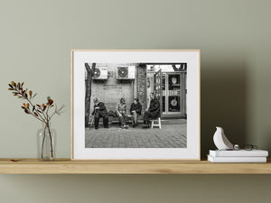 Two seated neighbours turn toward one another along a Beijing brick wall.