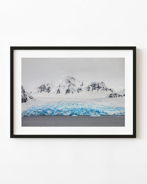 Blue glacier ice stretches beneath snowy Antarctic mountains and a grey sky.