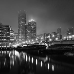 Illuminated Boston buildings rise beyond a bridge above dark water