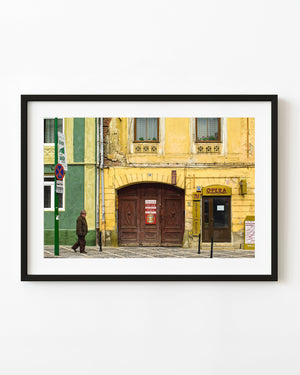 Brasov street photograph with a man walking past a warm yellow building and brown arched doors.