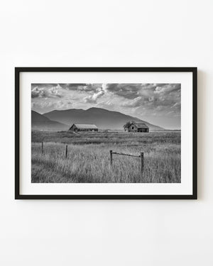 Farm buildings beneath mountains in British Columbia, photographed in black and white.