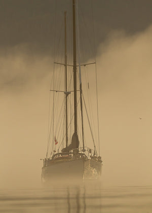 A two-masted sailboat and its reflection appear through warm morning fog.