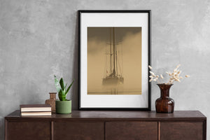 A small sailboat rests in muted gold and brown fog with a blurred reflection below.
