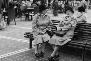 Older women sit side by side on a wooden bench beside outdoor café tables.