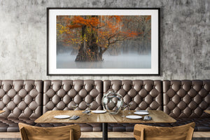 Ingelijst kunstwerk van een boom met herfstkleuren die boven een bruinleren kraampje in een restaurant hangt.