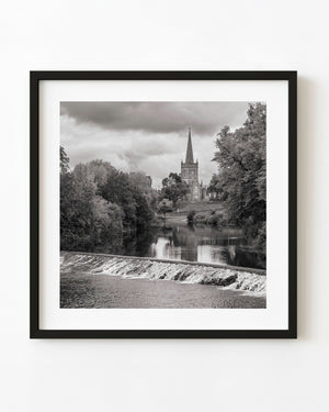 Black and white photograph of a Cahir church spire rising beyond a river weir and leafy trees.