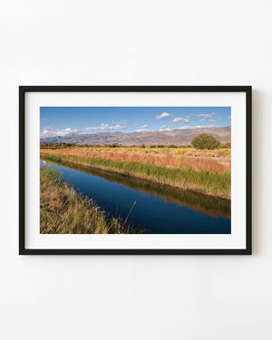A narrow blue river winds through golden grasses beneath distant California mountains and a clear sky.